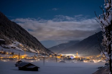 Avusturya 'nın Tirol şehri Neustift ve Pfarre kilisesine bakan kış gecesi manzarası dağlar ve bulutlar karşısında. Vadide sisli soğuk bir gece