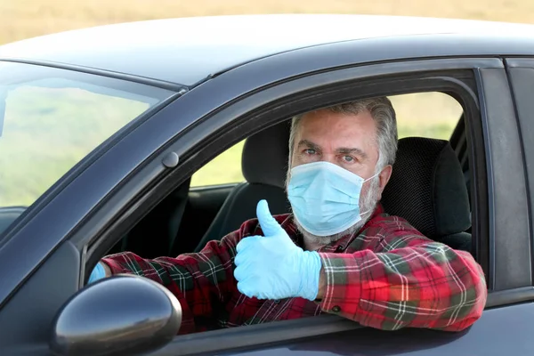 Adult driver with protective surgical mask and gloves gesturing with thumb up from car, corona virus protected person