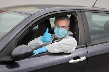 Koruyucu maske gözlüklü ve eldivenli yetişkin bir sürücü. Arabadan baş parmağıyla işaret ediyor. Corona virüsü korumalı biri.