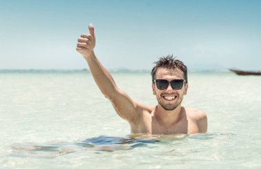 Güneş gözlüklü genç yakışıklı adam denizde eğleniyor ve baş parmağını kaldırıyor. Yaz tatilleri konsepti 