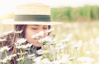 Şapkalı güzel genç kız güneşli bir günde vahşi papatyaların güzelliğinin ve kokusunun tadını çıkarıyor..