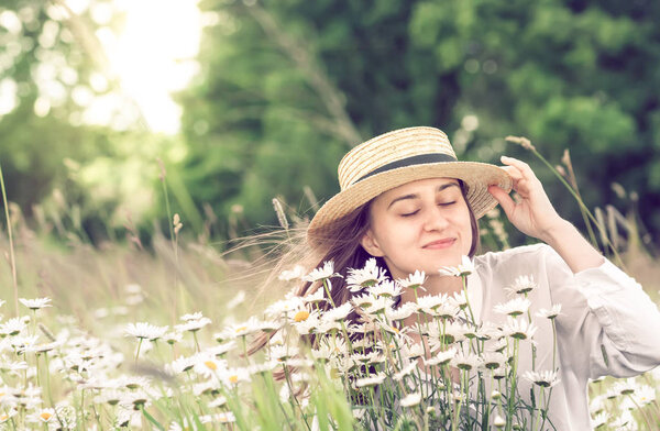 Beautiful young girl in a hat enjoys the beauty and smell of wild daisies on a warm sunny day.