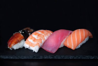 Japon klasik suşisi unagi, bataklık, ton balığı ve somonlu taş tabakta, siyah arka plan.