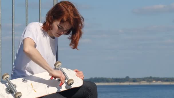 Das Mädchen repariert ein Skateboard, im Hintergrund ein Fluss. — Stockvideo