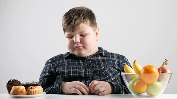 Young fat boy chooses between cakes and fruits 50 fps — Stock Video
