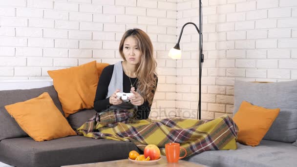 Traurige asiatische Mädchen sitzt auf der Couch unter der Decke und spielt das Konsolenspiel, verliert 50 fps — Stockvideo