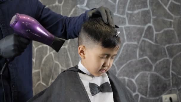 Парикмахер в черных перчатках сушит азиатского мальчика с феном 60 кадров в секунду — стоковое видео