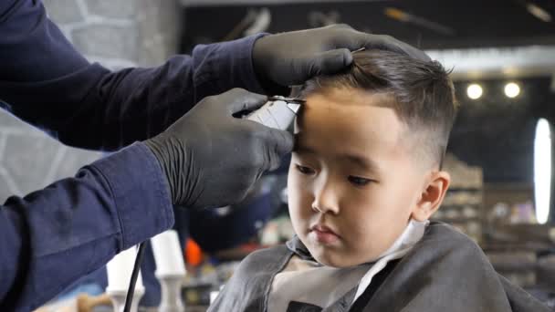 Fryzjer w czarne rękawiczki delikatnie goli grzywka smutny Asian dziecko 60 fps — Wideo stockowe