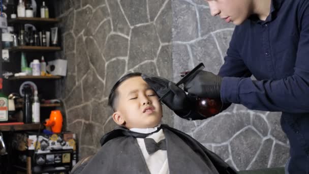 Парикмахер в черных перчатках распыляет и расчесывает волосы азиатского ребенка на 60 кадров в секунду — стоковое видео
