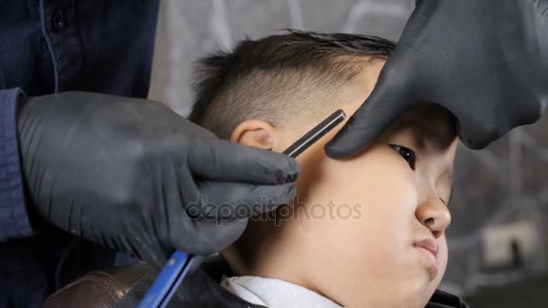 Peluquero con guantes negros afeita whisky con una navaja de afeitar a un niño asiático 60 fps — Vídeos de Stock