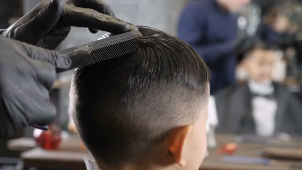 Peluquero con guantes negros peinando el pelo de un niño asiático y preparándose para cortarlos 60 fps — Vídeo de stock