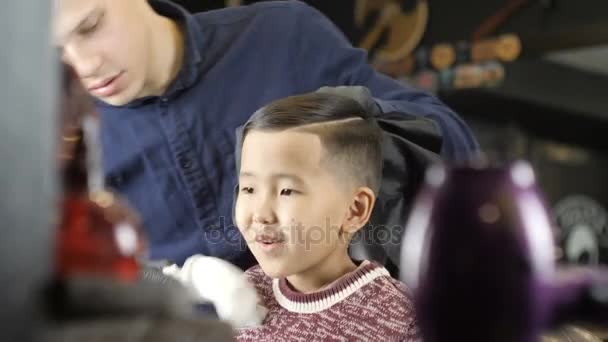 Парикмахер в черных перчатках делает прическу для азиатского ребенка, снимает отрезанные волосы с шеи кистью 60 кадров в секунду — стоковое видео