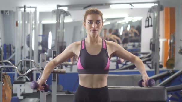 Deportiva joven haciendo lado mancuerna plantea mientras está sentado en el banco de deportes en un gimnasio. Vista lateral frontal 60 fps de cerca — Vídeos de Stock