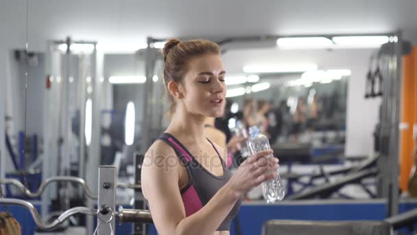 Deportiva joven bebe agua de una botella de plástico en el gimnasio entre ejercicios de 60 fps — Vídeo de stock