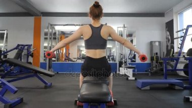 Spor genç kız tarafı dumbbell yapıyor bir spor salonunda spor bankta otururken yükseltir. Arka taraf görüntülemek 60 fps