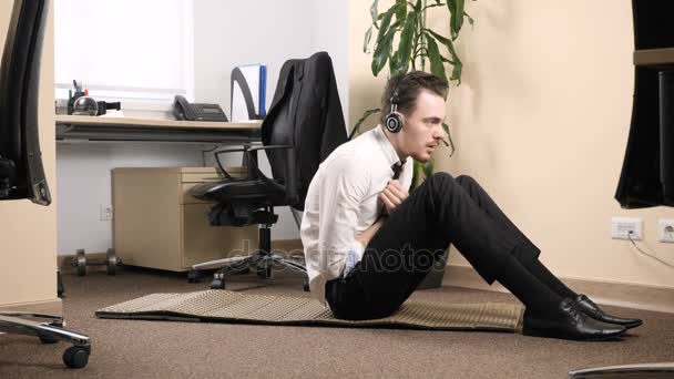 Genç adam bir takım elbise ve karın egzersizleri bir ofis ortasında gerçekleştirme giyen kulaklık 60 fps — Stok video