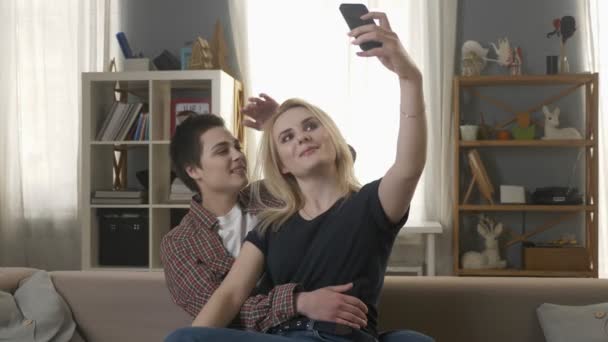 Dua gadis muda dan cantik duduk di sofa dan membuat selfie, lesbian, LGBT, pirang, berambut cokelat. 60 fps — Stok Video