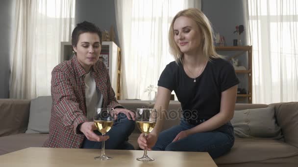 Two young, beautiful girls are sitting on the couch, raise glasses with wine and clinking, lesbians, LGBT, blonde, brunette 60 fps — Stock Video