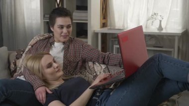 60 fps gülüyor bir esmer ve kullanarak kırmızı laptop, rahatlık, kucaklamaları, ekose, kucağına genç sarışın bir kadın yatıyor