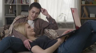 Genç sarışın bir kadın yatıyor tur kanepede ve kullanma kırmızı laptop, rahatlık, kucaklamaları, ekose, konuşurken, lezbiyenler, lgbt, tartışma bir esmer 60 fps