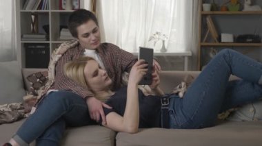 Lezbiyen çift kanepede tablet bilgisayar, tablet, 60 fps gülümseyerek elele fotoğraf kaydırma kullanarak dinleniyor