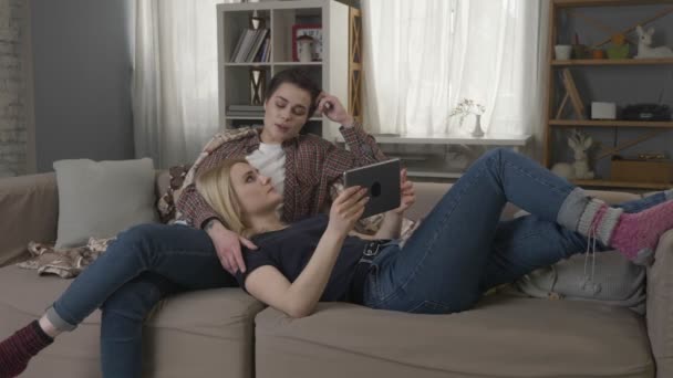 Лесбиянки отдыхают на диване, используя планшетный компьютер, девушка с короткими волосами нежно гладит шею своих партнеров 60 fps — стоковое видео