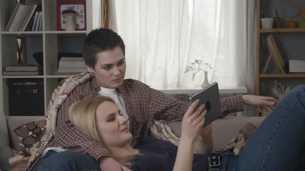 Lesbický pár se odpočinku na gauči, používající počítač tabletu, posouvání fotografie na tabletu, držení rukou, usmívající se, mluví, rodinné idyly, láska, roztomilý 60 fps — Stock video