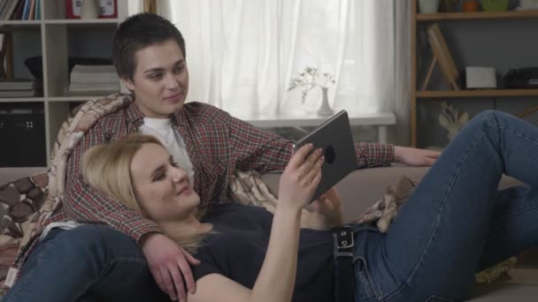Lesbický pár sedí na gauči, pomocí tabletového počítače, posouvání fotek na tabletu, držení rukou, s úsměvem, mluvící, rodinné idyly se 60 fps — Stock video