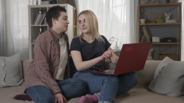 Två unga lesbisk tjejer är sitter i soffan, med en dator, rullning internet, shopping online, ler, pratar 60 fps — Stockvideo