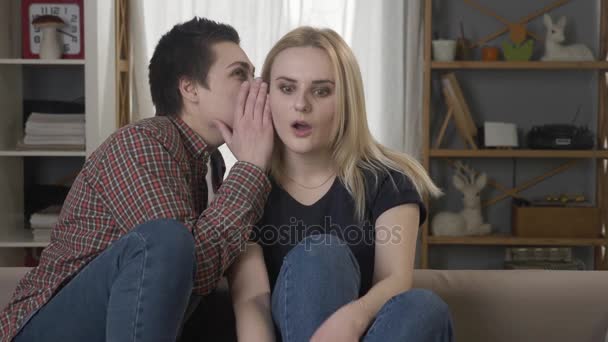 Две молодые лесбиянки сидят на диване, девушка с короткими волосами говорит секрета своему партнеру, блондинка шокирована 60 кадров в секунду — стоковое видео