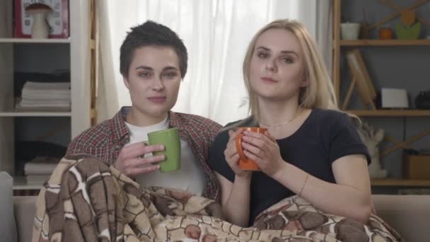 Two young lesbian girls are sitting on the couch, covered with a warm blanket, holding cups in their hands, drinking dark tea, coffee, cuddling, looking at the camera 60 fps — Stock Video