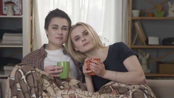 Two young lesbian girls are sitting on the couch, covered with a warm blanket, holding cups in their hands, cuddling, looking at the camera 60 fps — Stock Video