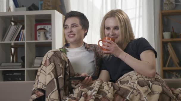 Duas jovens lésbicas estão sentadas no sofá, cobertas com um cobertor quente, segurando copos em suas mãos, bebendo chá escuro, café, abraçando, assistindo TV, trocando de canais 60 fps — Vídeo de Stock
