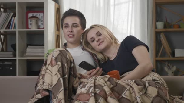 Dos chicas lesbianas jóvenes están sentadas en el sofá, cubiertas con una manta caliente, sosteniendo tazas en sus manos, bebiendo té oscuro, café, abrazos, viendo la televisión, cambiando de canal, riendo 60 fps — Vídeo de stock