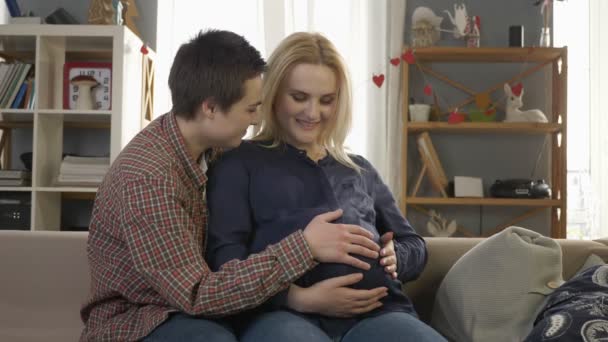 Dwie młode dziewczyny lesbijki, siedząc na kanapie, w ciąży Blondynka pieści brzuch, czekając na wygląd babys, przytulności, miłości, szczęścia, kissing.60 fps — Wideo stockowe