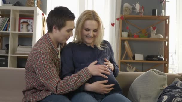 Zwei junge lesbische Mädchen sitzen auf der Couch, schwangere Blondine streichelt den Bauch, wartet auf die Babys Aussehen, Gemütlichkeit, Liebe, Glück, küssen 60 fps — Stockvideo