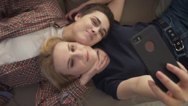 Dos chicas jóvenes yacen en el sofá, hacer selfie en un teléfono inteligente, mueca, hacer caras divertidas, top shot 60 fps — Vídeos de Stock