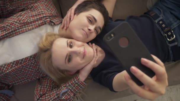 Две молодые девушки лежат на диване, делают селфи на смартфоне, улыбаются, влюбленные, lga, молодая пара, топ-снимок 60 кадров в секунду — стоковое видео