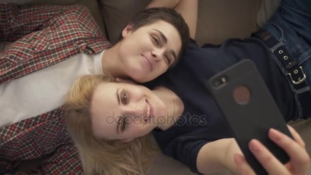 Две юные лесбиянки лежат на диване, делают селфи на смартфоне, любовники, неженки, молодая пара. верхний снимок 60 кадров в секунду — стоковое видео
