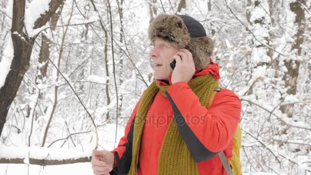 Anciano europeo hablando en un smartphone en un bosque nevado. Grueso matorral denso de árboles y raíces en el bosque cubierto de nieve. Caminata y concepto de viaje 60 fps — Vídeo de stock