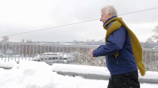 Um homem europeu idoso corre uma cruz em uma estrada nevada, inverno, esportes, cardio. Conceito de vida saudável. 60 fps — Vídeo de Stock