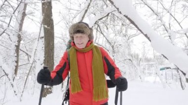 Yaşlı beyaz gülümseyen adam Hiking sopa ile karlı bir orman yoluyla girer. Portre video çekim. Ağaçlar karla kaplı ormanda kalın yoğun çalılık. Yürüyüş ve kavram seyahat 60 fps.
