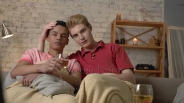 Çok uluslu gay çift sıcak bir battaniye ile örtülü kanepede oturup Tv izlemek, uzaktan kumandayı, kameraya bak. Homeliness, romantik akşam, arka plan, hugs, mutlu Lgbt aile kavramı