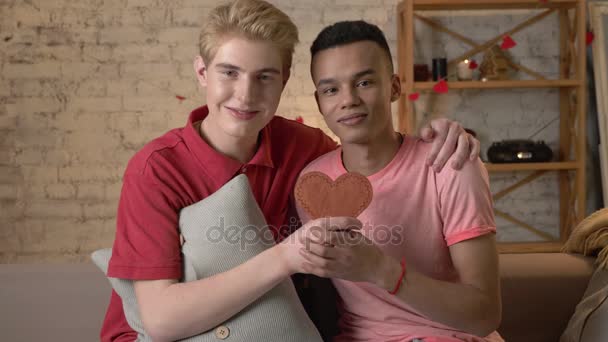 Mnohonárodnostní šťastné gay pár sedí na gauči a drží srdce se v rukou. Nefalšovaná, Lgbt loverhappy s, šťastné gay koncepce. 60 snímků za sekundu — Stock video