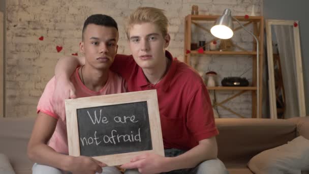 Un triste couple gay international est assis sur le canapé et tient une pancarte. Nous n'avons pas peur. Regarde la caméra. Confort à la maison sur le fond. 60 ips — Video