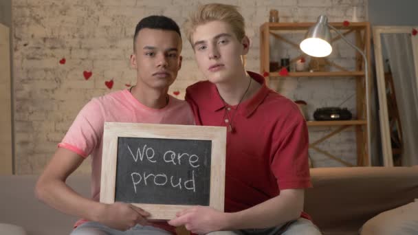 Грустная международная гей-пара сидит на диване и держит табличку. Мы гордимся. Посмотри в камеру. Домашний комфорт на заднем плане. 60 кадров в секунду — стоковое видео