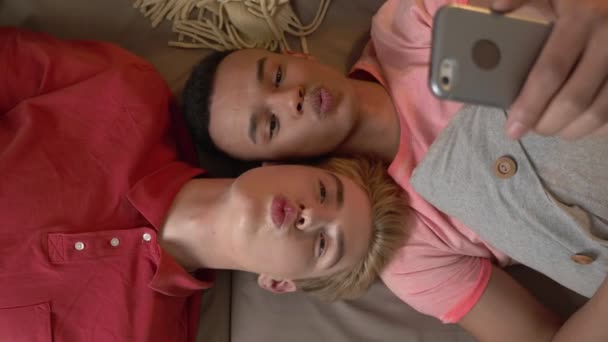 Международная гей-пара лежит на диване и делает селфи на смартфоне. Гомосексуализм, ЛГБТ-ловер Счастливая семейная концепция. Топ выстрел 60 кадров в секунду — стоковое видео