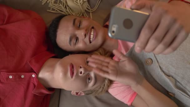 Feliz casal gay internacional está deitado no sofá e fazendo selfies em um smartphone. Homeliness, amantes LGBT, conceito de família gay feliz, rindo. Diz hallo. Conversa de vídeo. Tiro superior 60 fps — Vídeo de Stock