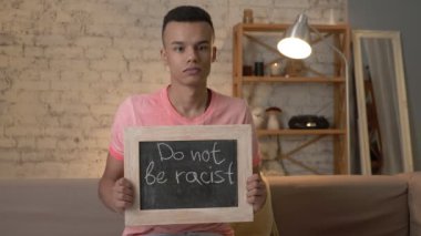 Üzgün bir Afrika adam kanepede oturur ve bir işaret tutar. Irkçı olmayın. Anti-ırkçılık, ulusal eşitsizlik kavramı. Kameraya bak. 60 fps