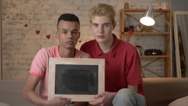 Una triste coppia gay internazionale è seduta sul divano con un cartello vuoto. Comfort domestico sullo sfondo. 60 fps — Video Stock